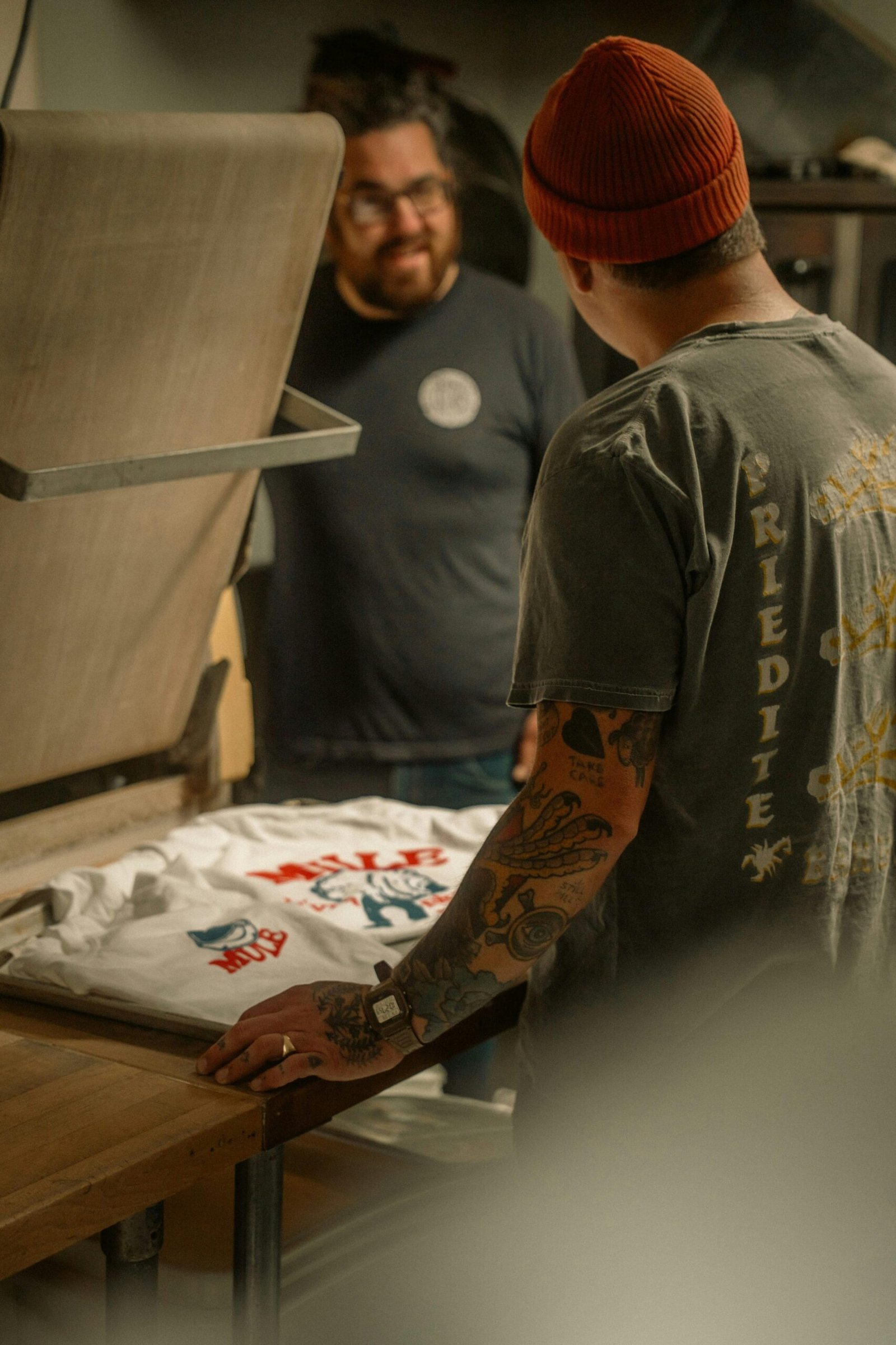 Two men engaged in screen printing a t-shirt in a cozy workshop setting.